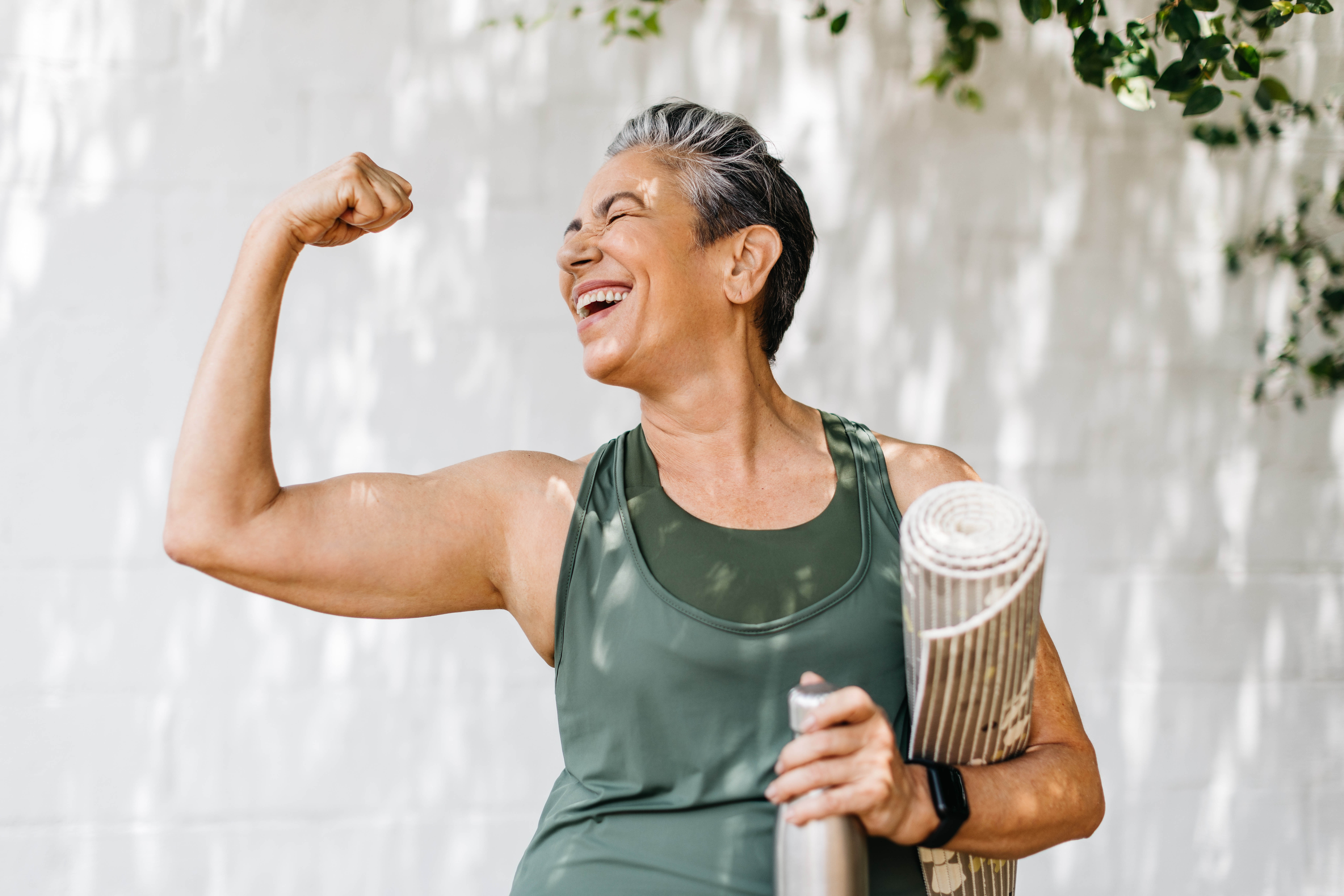 Older woman flexing her arm and smiling, she has a yoga mat under her other arm Older woman flexing her arm and smiling, she has a yoga mat under her other arm